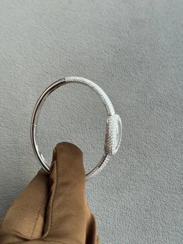 Close-up of a silver, textured bracelet being held by a brown glove, against a light gray textured background, highlighting the ring's sparkling surface.
