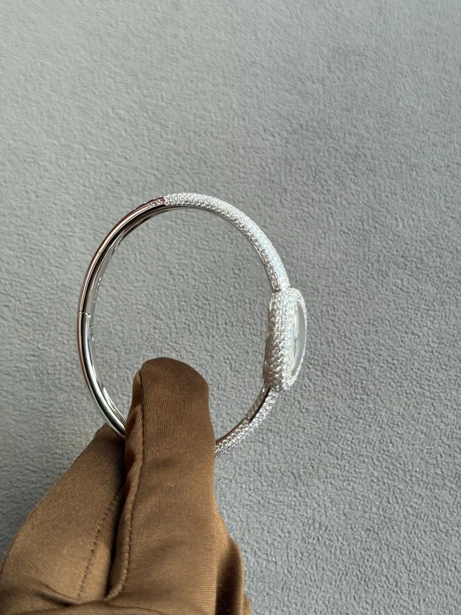 Close-up of a silver, textured bracelet being held by a brown glove, against a light gray textured background, highlighting the ring's sparkling surface.