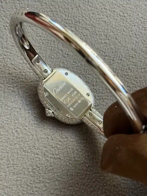 Close-up of a silver Cartier wristwatch with an open deployment clasp, showing engraved back and brown strap on a gray fabric surface.
