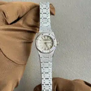 Close-up of a glittery silver Audemars Piguet watch with a textured dial, held by a gloved hand against a gray backdrop.