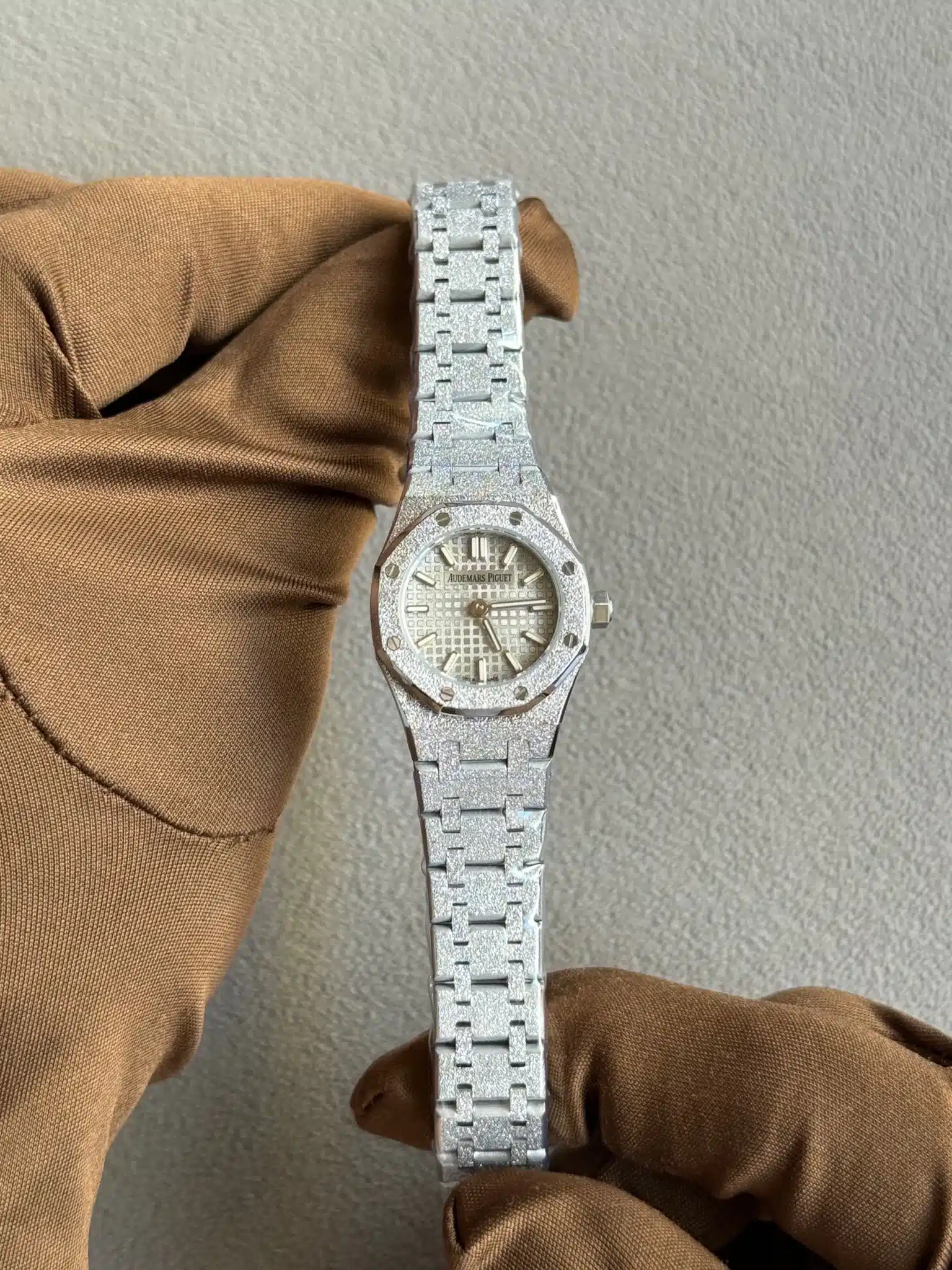 Close-up of a glittery silver Audemars Piguet watch with a textured dial, held by a gloved hand against a gray backdrop.