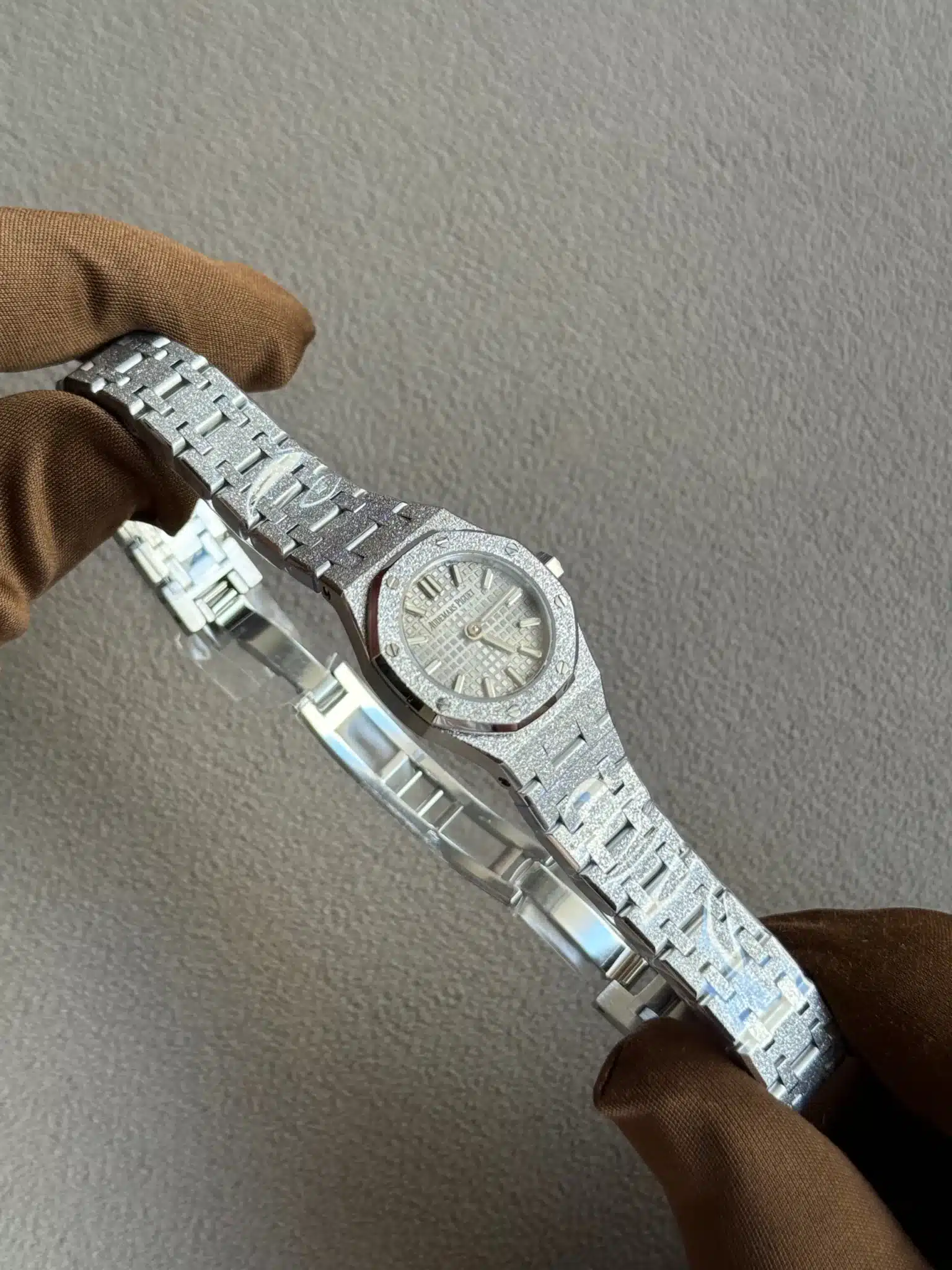 Gloved hand holding a silver textured stainless steel wristwatch with a patterned dial against a gray background.
