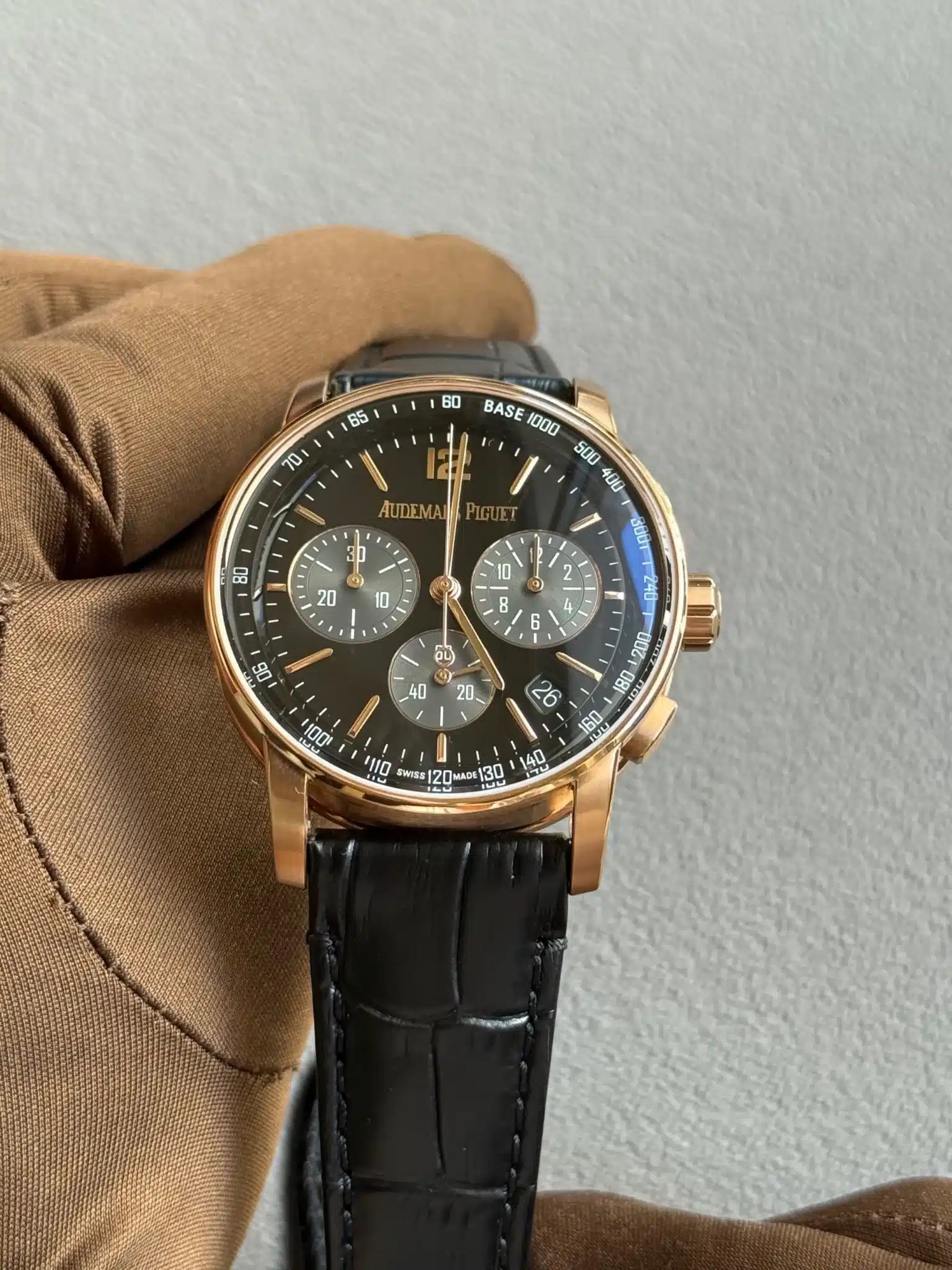 Gold Audemars Piguet chronograph watch with black dial and multiple subdials, held by a gloved hand.