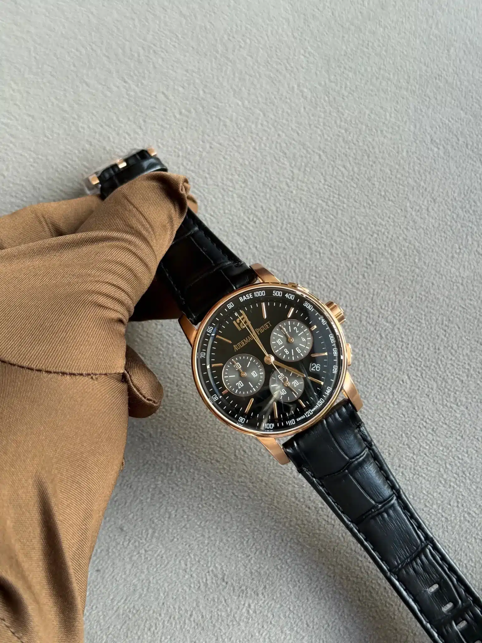 Gold-tone chronograph watch with black dial on a black leather strap, worn on a brown-sleeved arm.