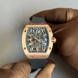 Rose gold Richard Mille watch with a skeleton dial held by a gloved hand against a gray background, black strap visible at the bottom.