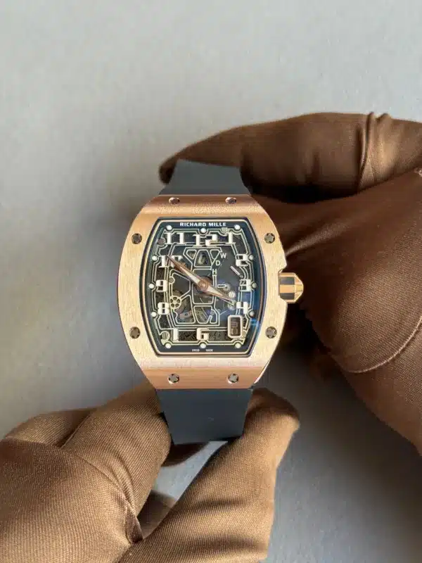 Rose gold Richard Mille watch with a skeleton dial held by a gloved hand against a gray background, black strap visible at the bottom.