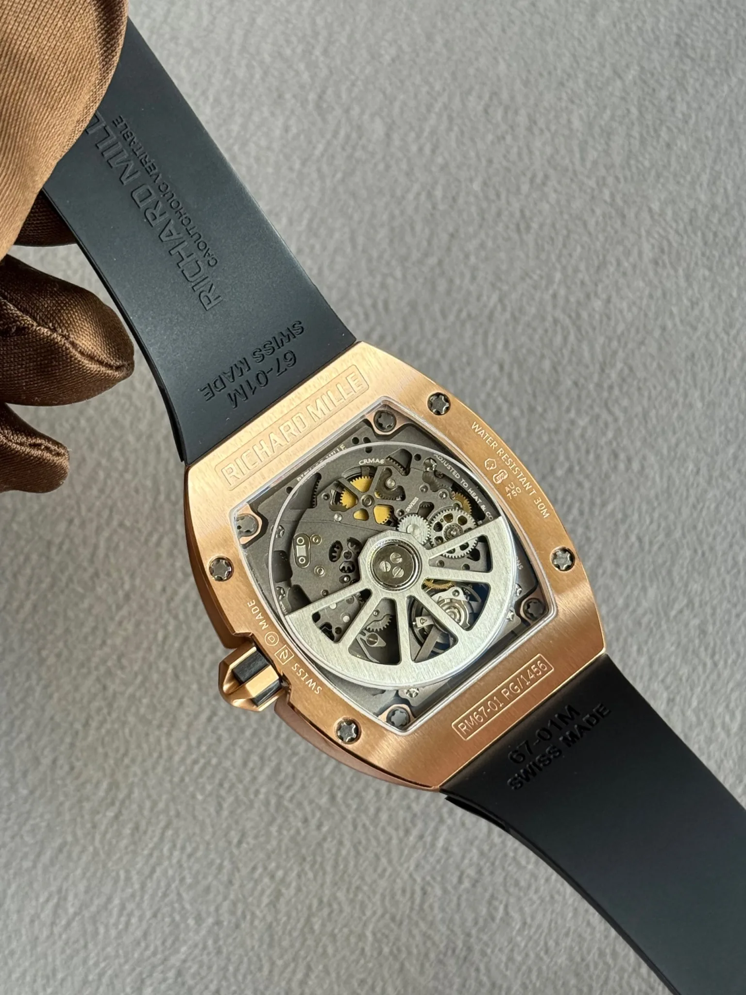 Close-up of a rose-gold Richard Mille wristwatch back with visible skeleton movement, black strap, held by a gloved hand.