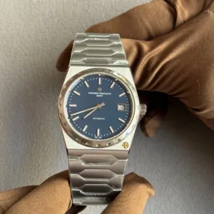 Vacheron Constantin Genève automatic wristwatch with a blue dial, date window at 3 o'clock, and stainless steel bracelet held by gloved hands.