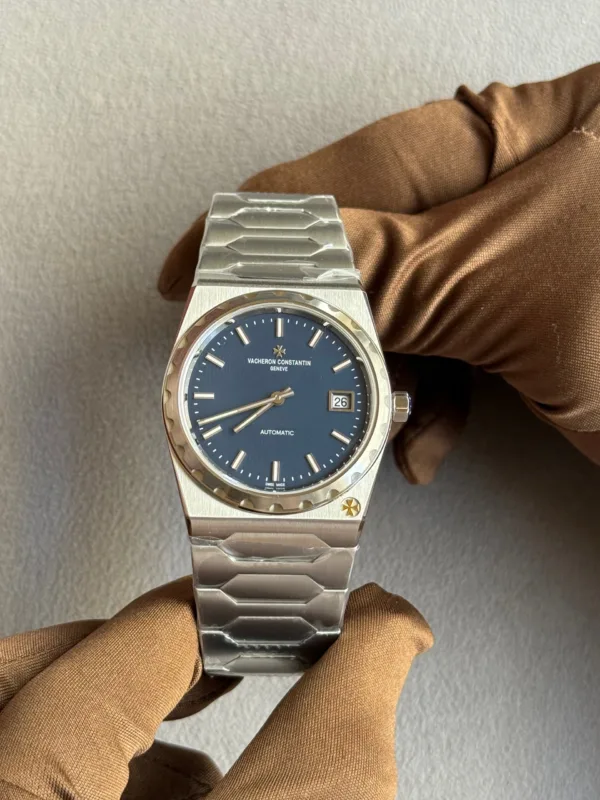 Vacheron Constantin Genève automatic wristwatch with a blue dial, date window at 3 o'clock, and stainless steel bracelet held by gloved hands.