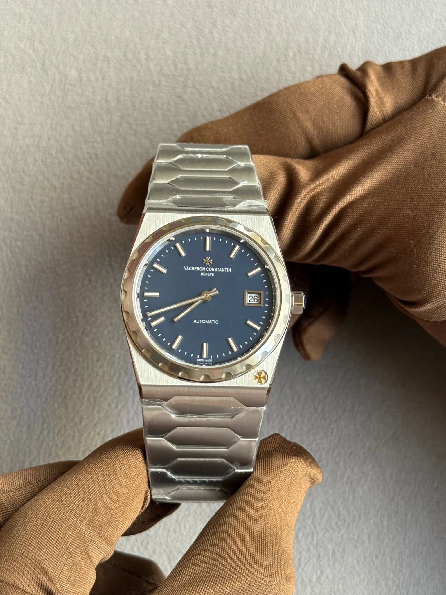 Vacheron Constantin Genève automatic wristwatch with a blue dial, date window at 3 o'clock, and stainless steel bracelet held by gloved hands.