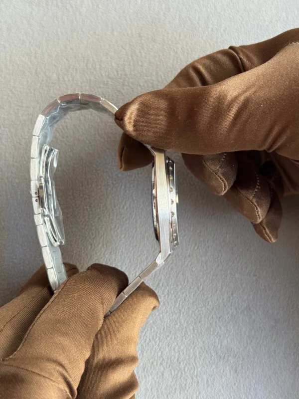 Person wearing brown gloves holding a silver stainless-steel watch by its bracelet, side view against a gray background.