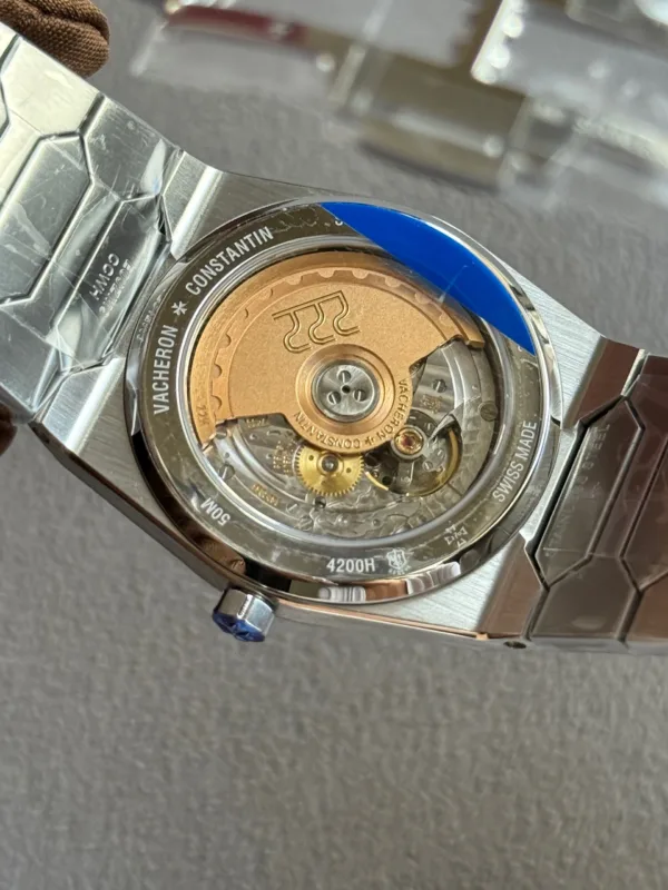 Close-up of a stainless steel watch case back with an exposed gold rotor, engravings read Vacheron Constantin and Swiss Made, blue protective sticker partially peeled.