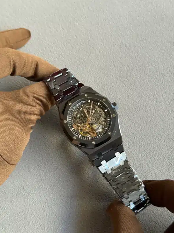 Close-up of a black skeleton wristwatch with visible gears on a metal bracelet, held by a gloved hand.
