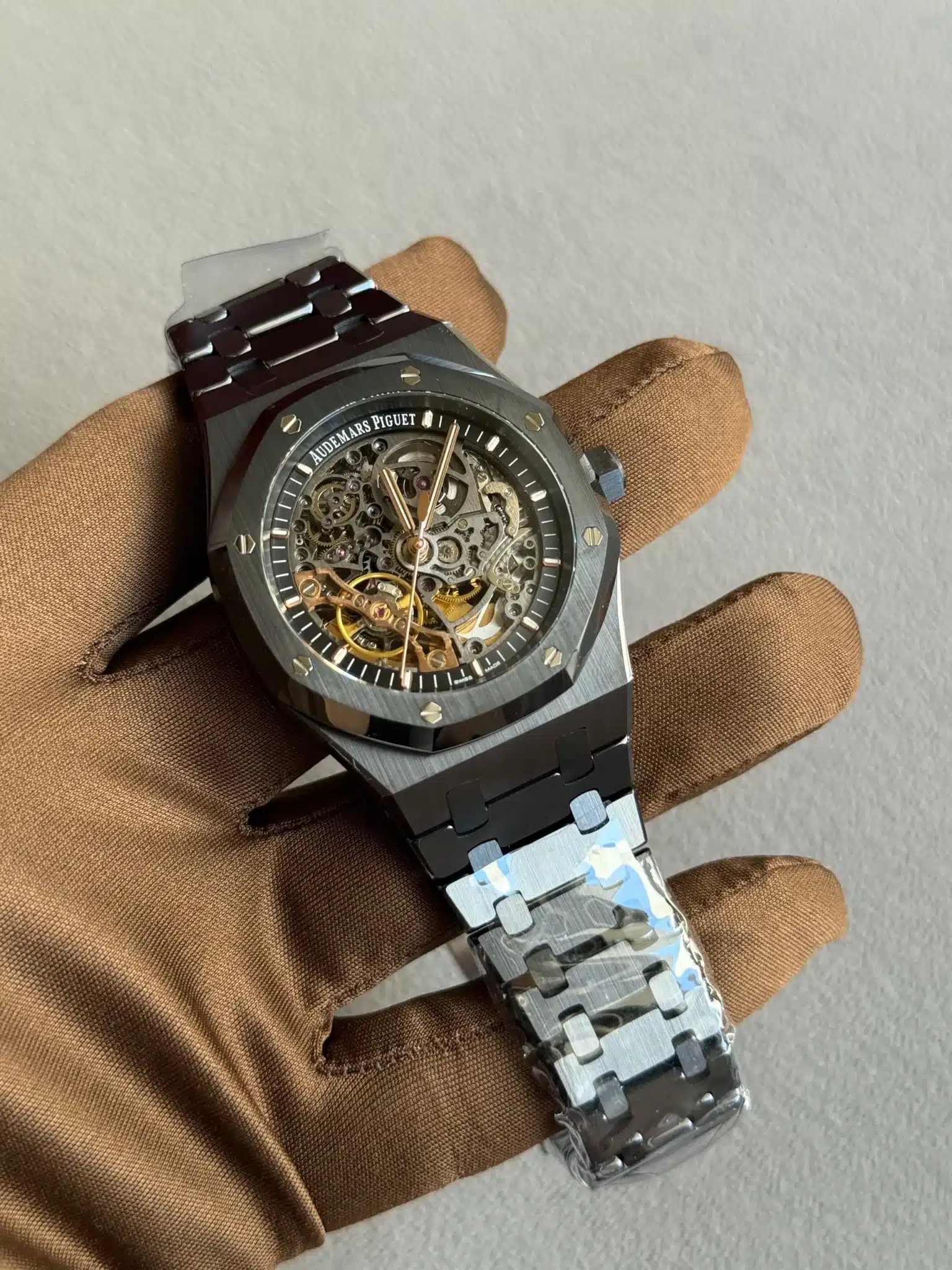 Skeleton Audemars Piguet watch with exposed gears held by a gloved hand on a gray backdrop, shows intricate inner mechanism and metallic strap
