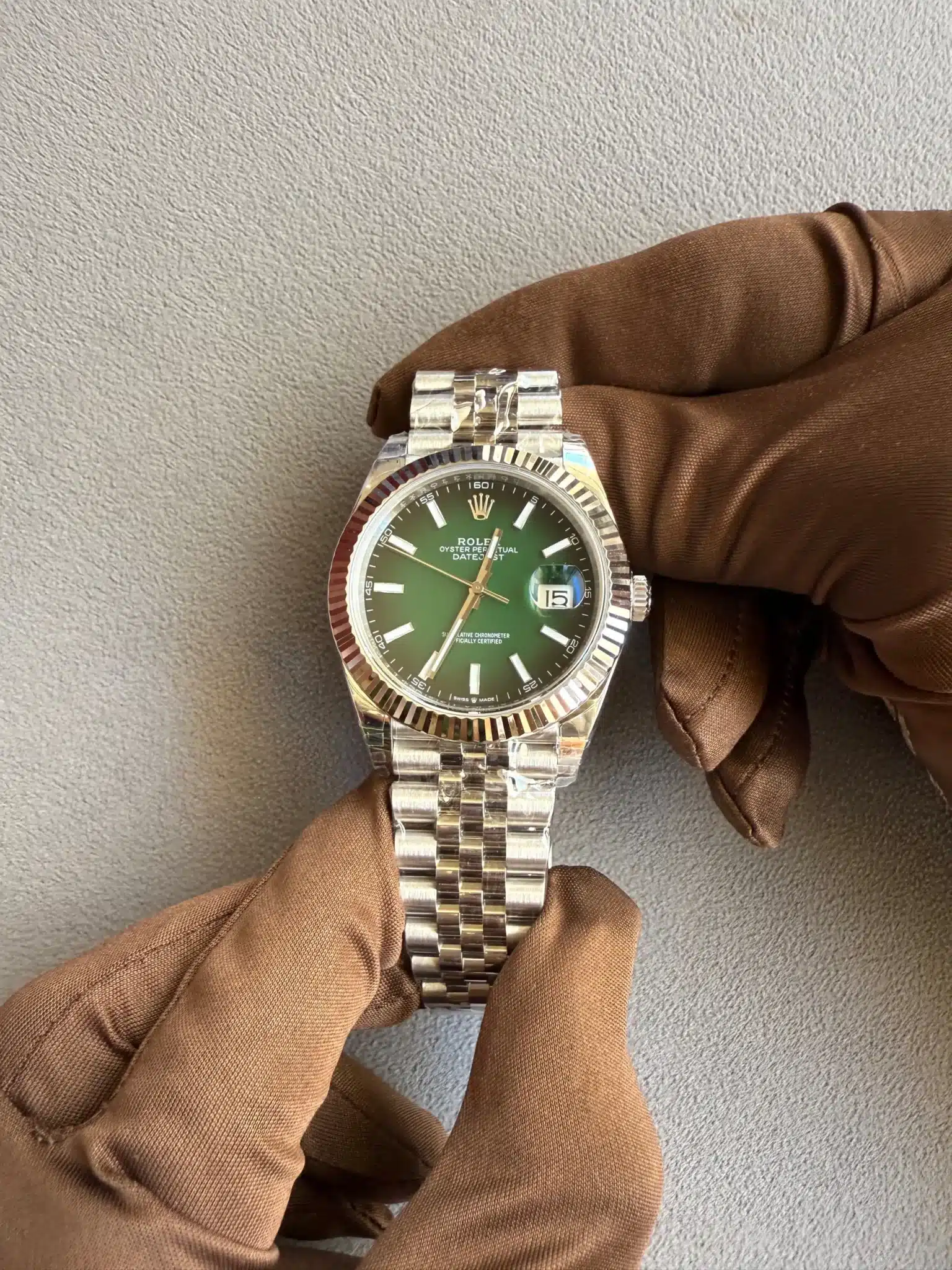 Rolex Datejust watch with a green dial and stainless steel bracelet held by brown-gloved hands against a gray textured background.