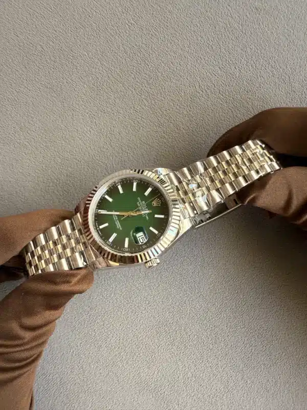 Stainless steel Rolex watch with a green dial being held by gloved hands against a textured gray background.
