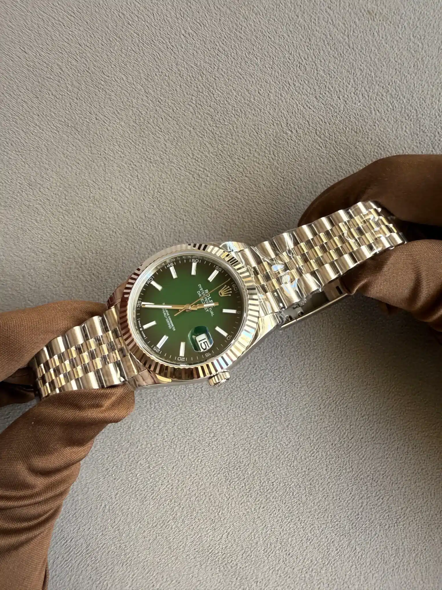 Stainless steel Rolex watch with a green dial being held by gloved hands against a textured gray background.