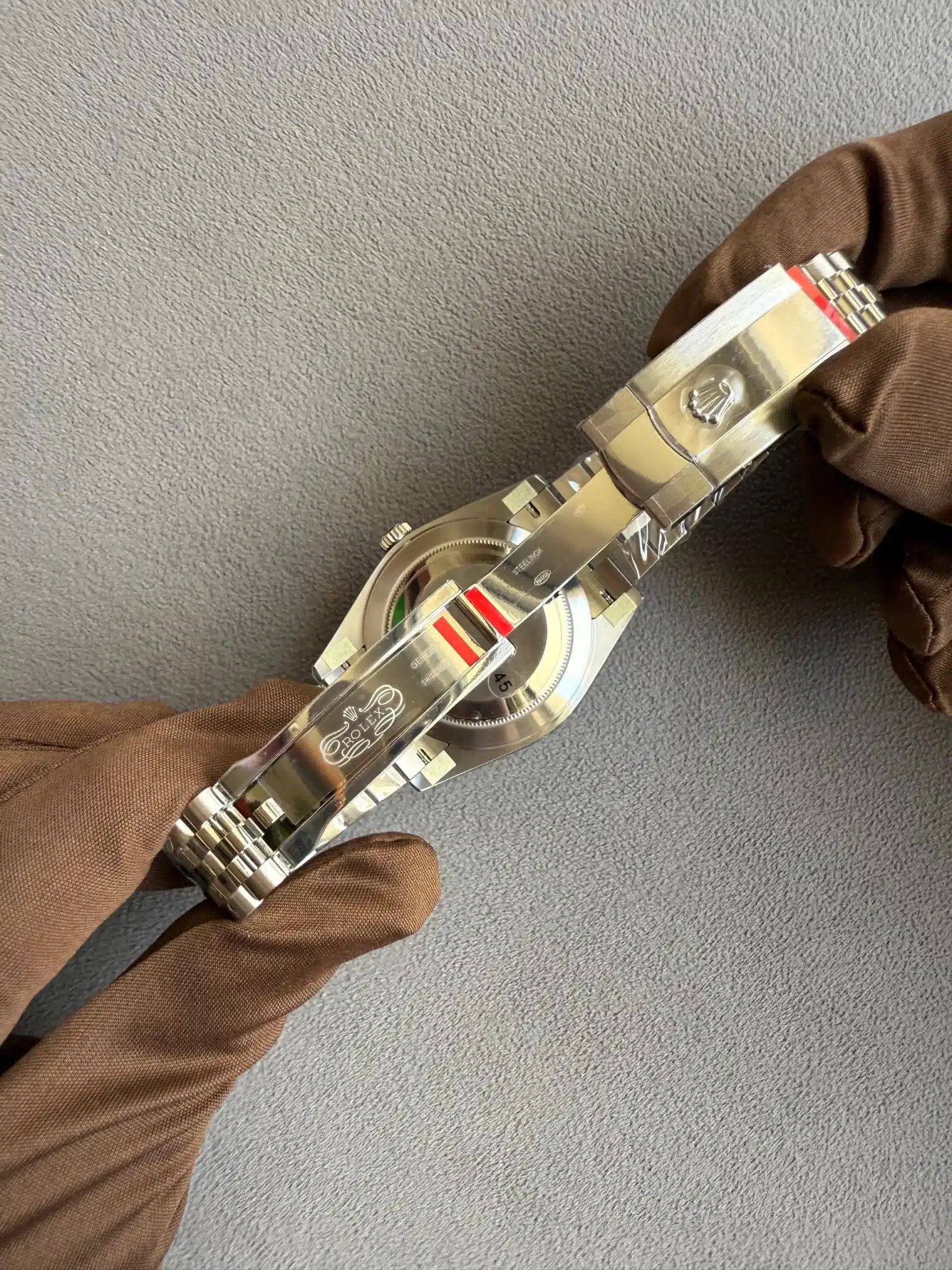 Close-up of a stainless steel Rolex watch held by gloved hands against a gray textured surface.