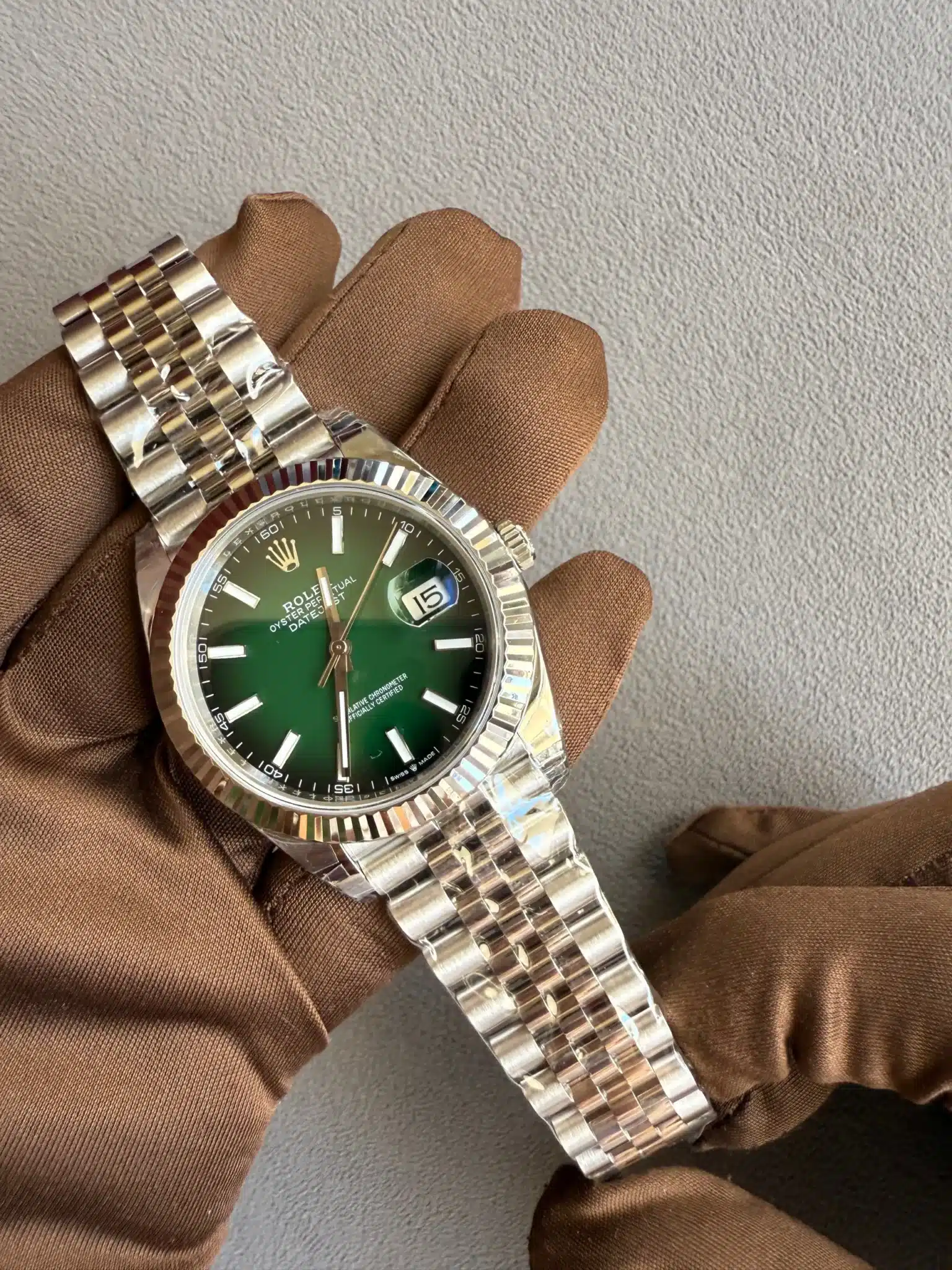 Rolex Datejust with a green dial and stainless steel jubilee bracelet, held by a brown-gloved hand.
