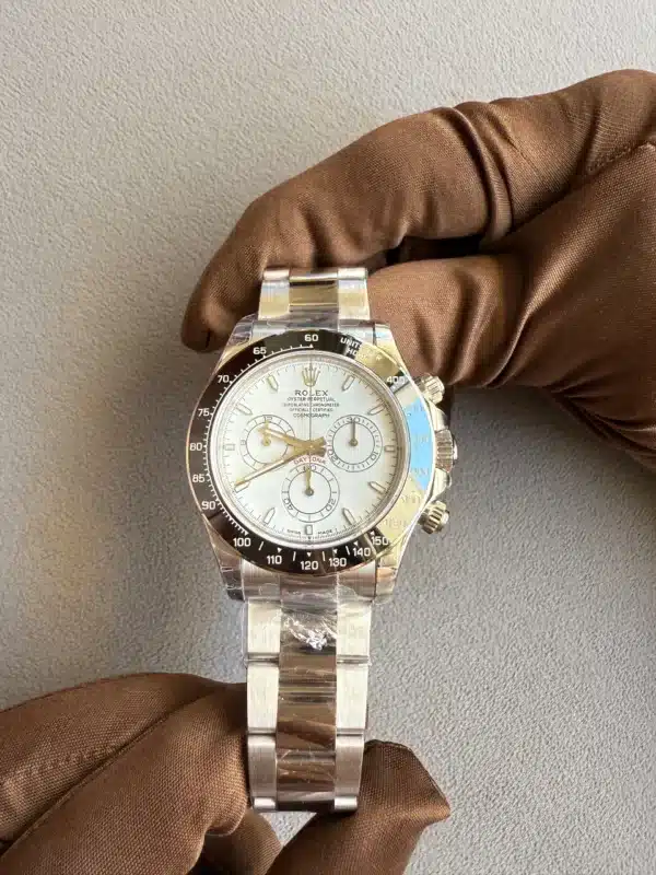 Close-up of a stainless steel Rolex Daytona chronograph being held by a brown-gloved hand against a gray textured background for display purposes.