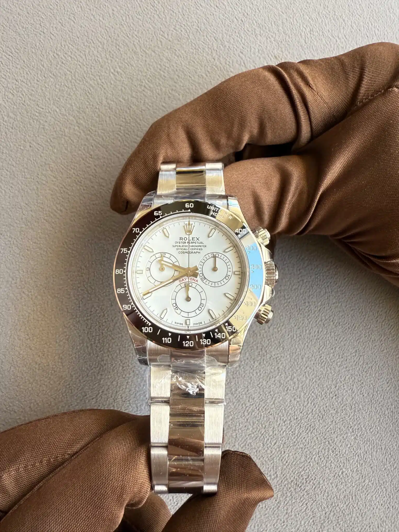 Close-up of a stainless steel Rolex Daytona chronograph being held by a brown-gloved hand against a gray textured background for display purposes.