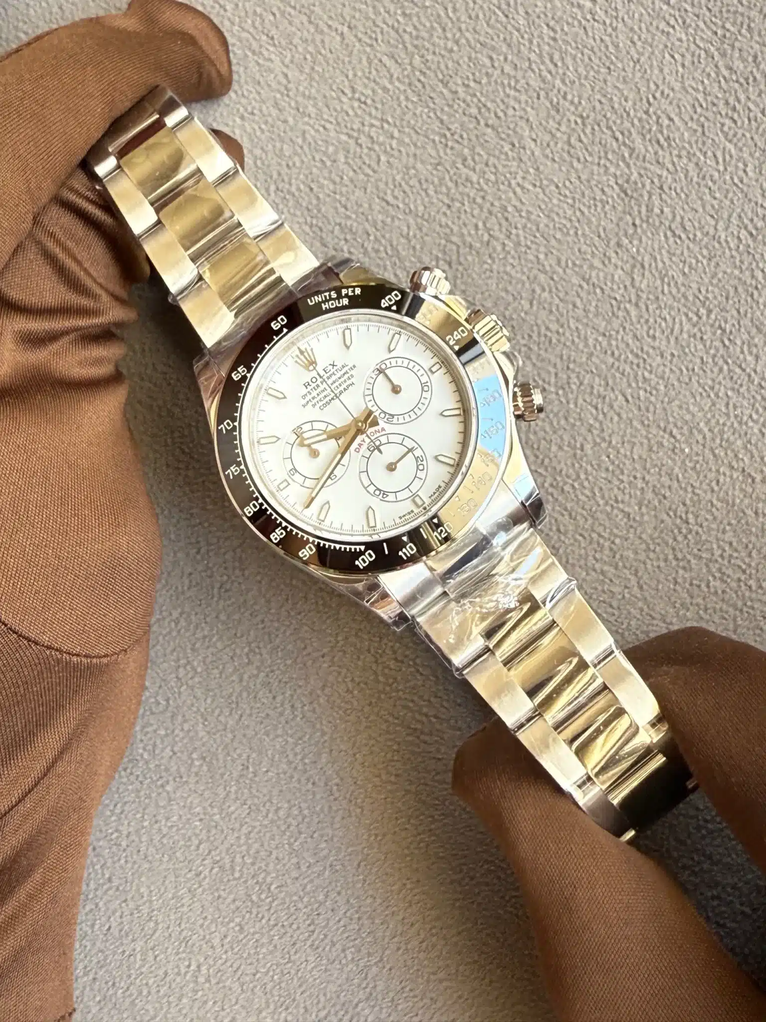 Close-up of a silver Rolex wristwatch with a white dial and black bezel, resting on a brown-gloved hand against a gray background.