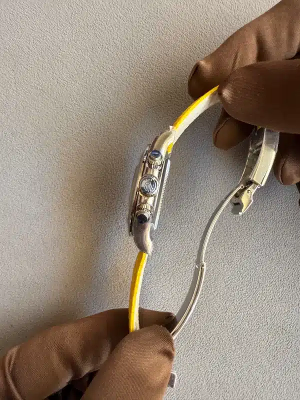 Close-up of a gloved hand using a precision tool to adjust a metallic wristwatch with a yellow strap on a gray surface, viewed from the side.