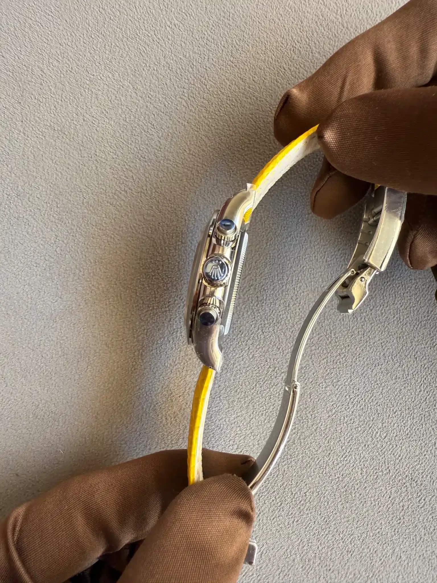 Close-up of a gloved hand using a precision tool to adjust a metallic wristwatch with a yellow strap on a gray surface, viewed from the side.