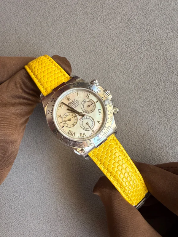 Close-up of a silver Rolex watch with a cream dial and yellow textured strap held by a gloved hand against a gray surface.