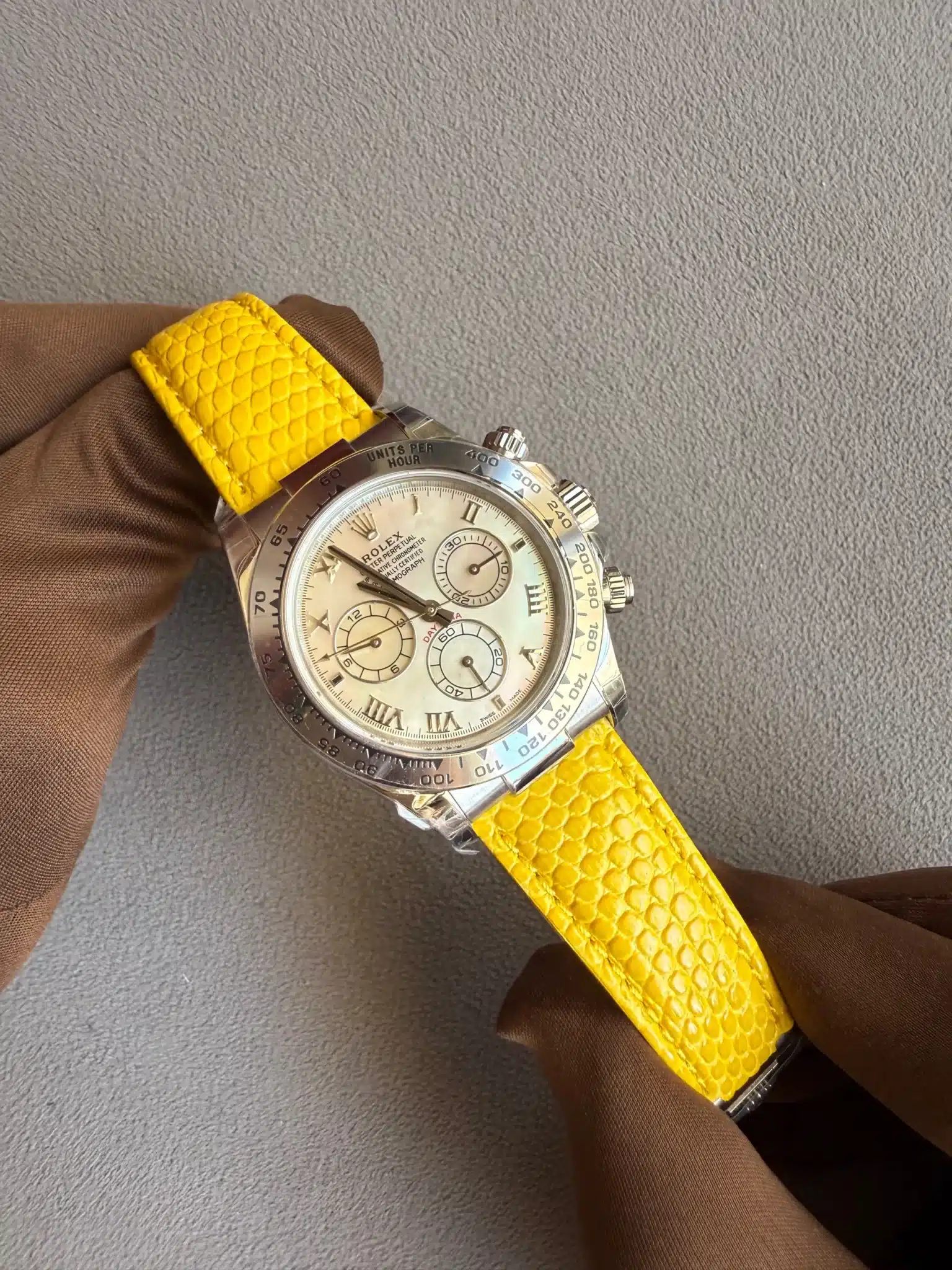 Close-up of a silver Rolex watch with a cream dial and yellow textured strap held by a gloved hand against a gray surface.