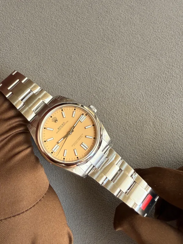 Close-up of a stainless steel Rolex Oyster Perpetual wristwatch with a peach dial on a brown sleeve, resting on a gray background.