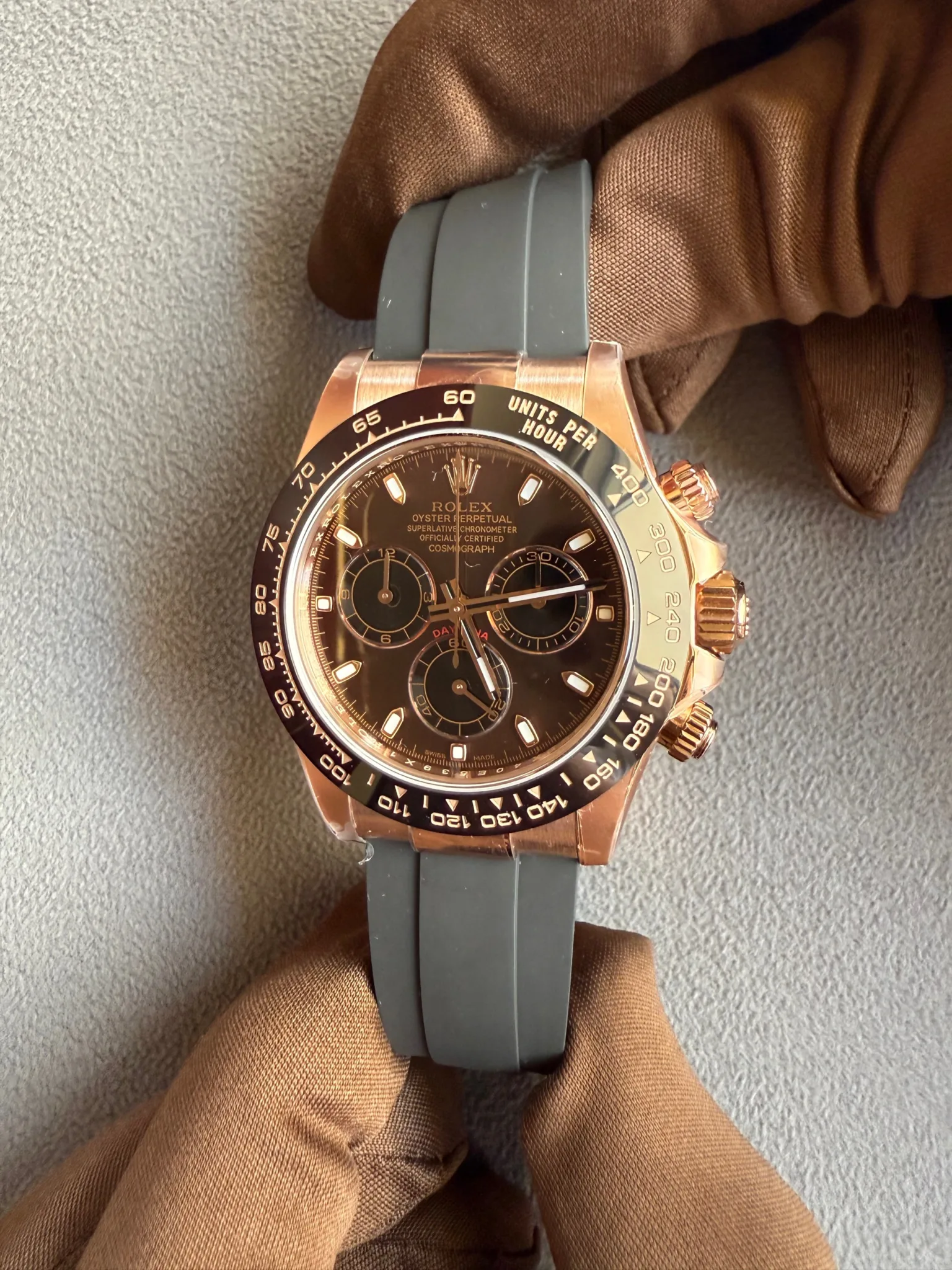 Rose gold Rolex Daytona with black dial and tachymeter bezel, on a gray rubber strap, held by brown gloves.