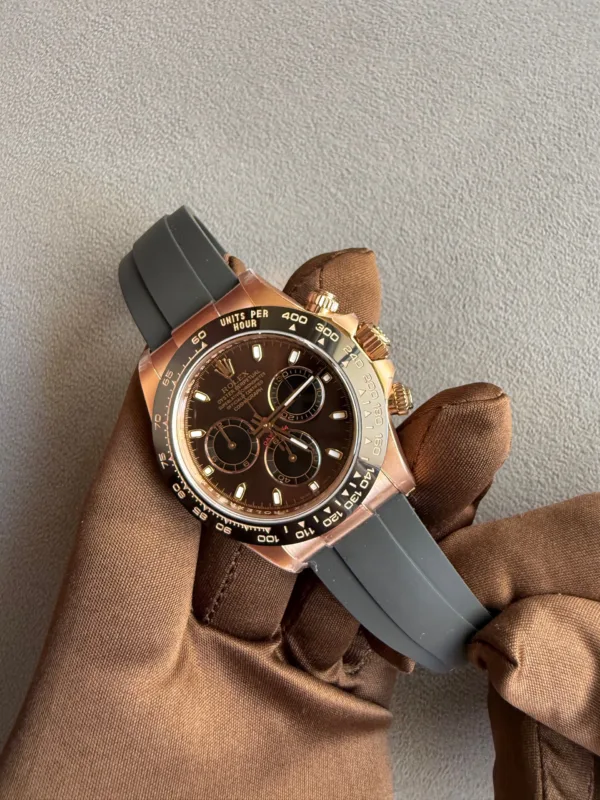 Rose gold Rolex chronograph with a black dial, gray rubber strap, held by brown-gloved hand against a textured gray background.
