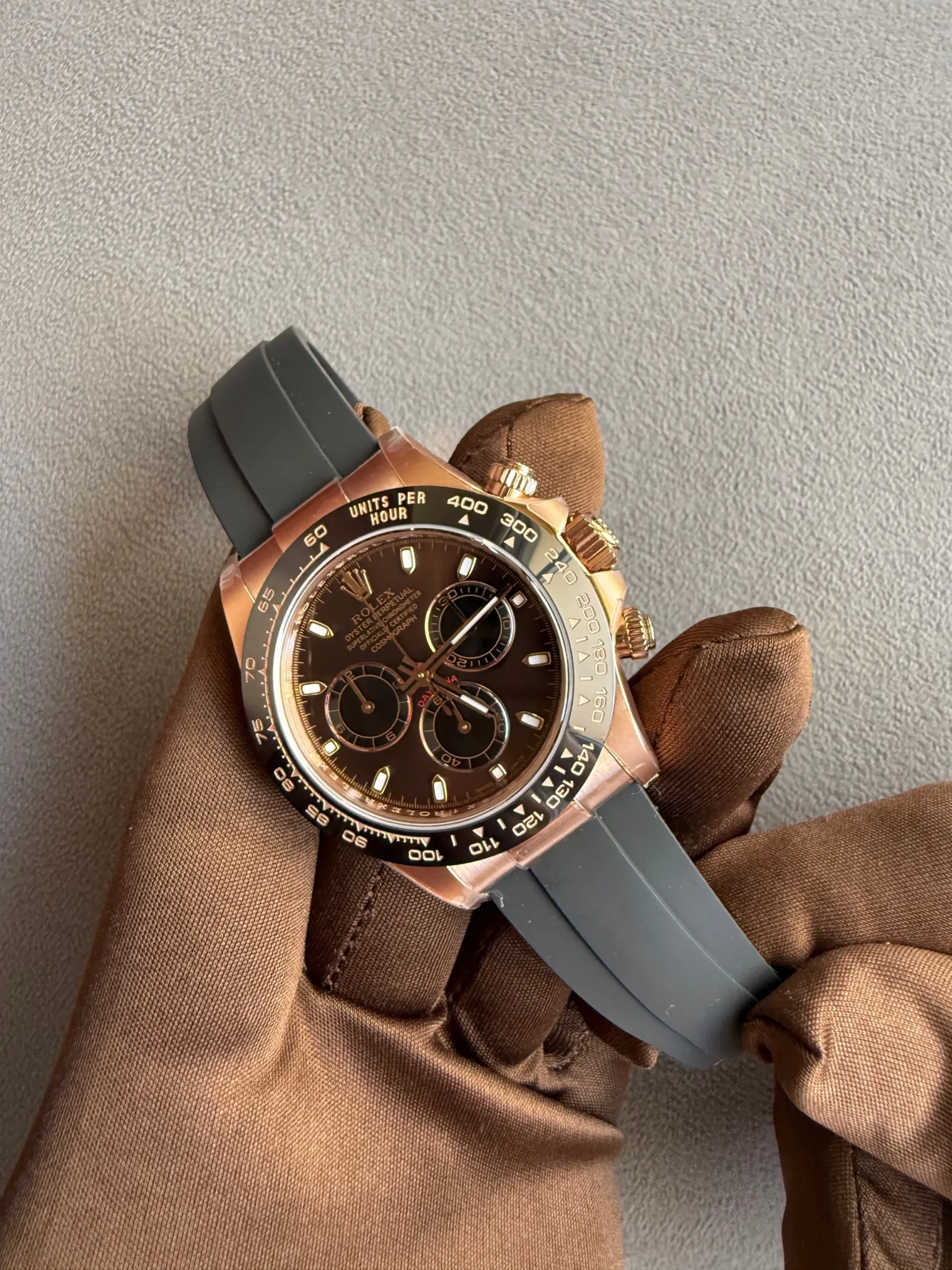 Rose gold Rolex chronograph with a black dial, gray rubber strap, held by brown-gloved hand against a textured gray background.