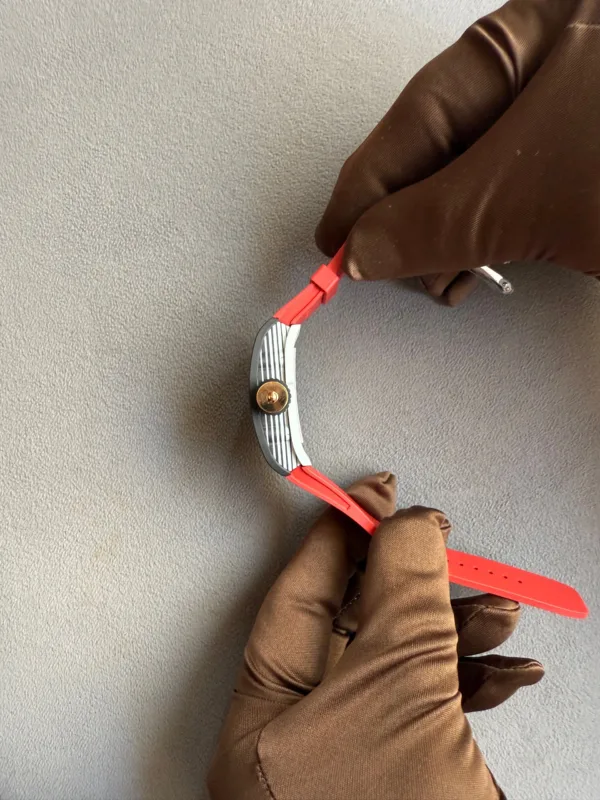 Two gloved hands connect a red watch strap to a metallic watch case against a gray textured background.