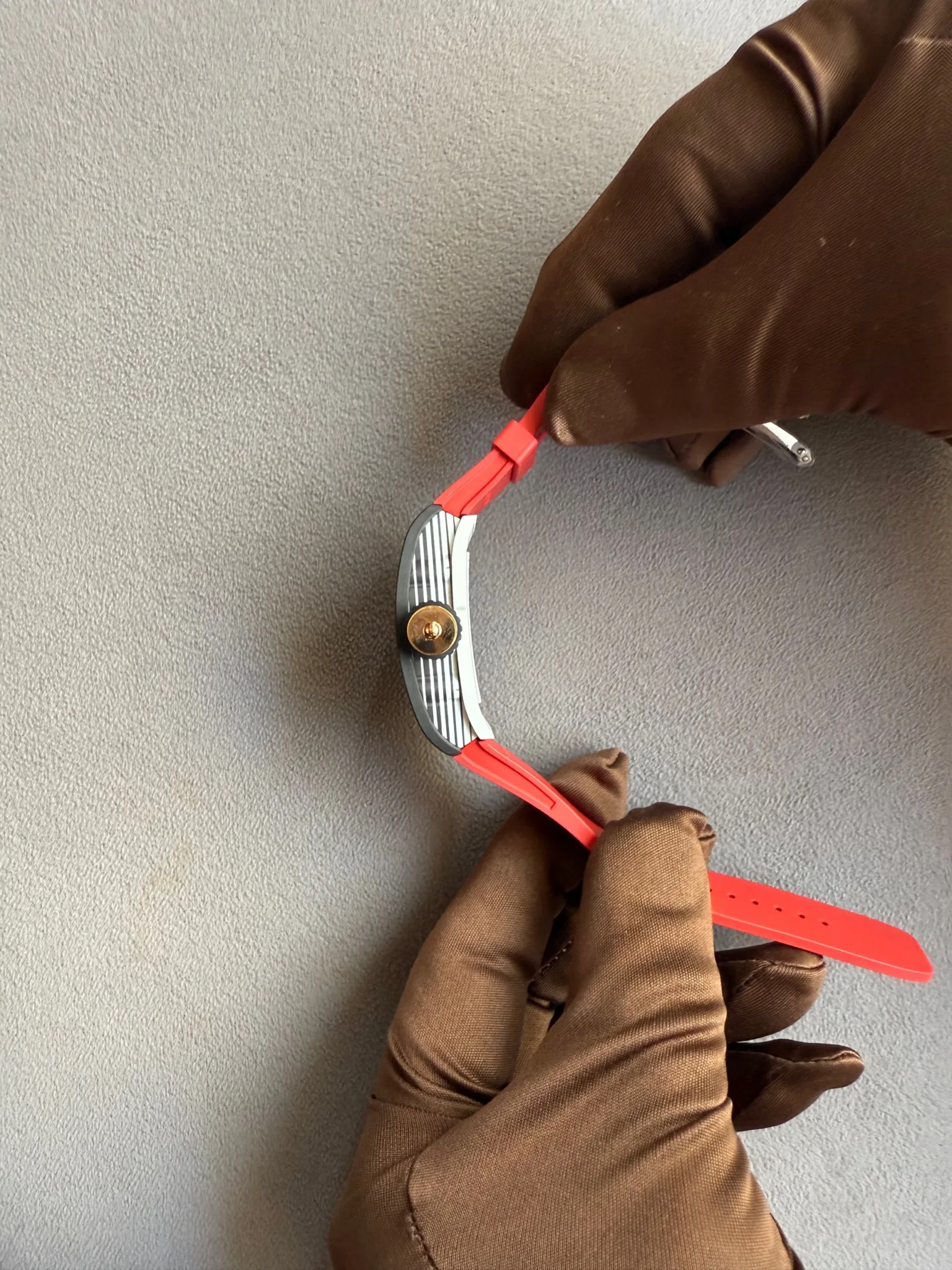 Two gloved hands connect a red watch strap to a metallic watch case against a gray textured background.