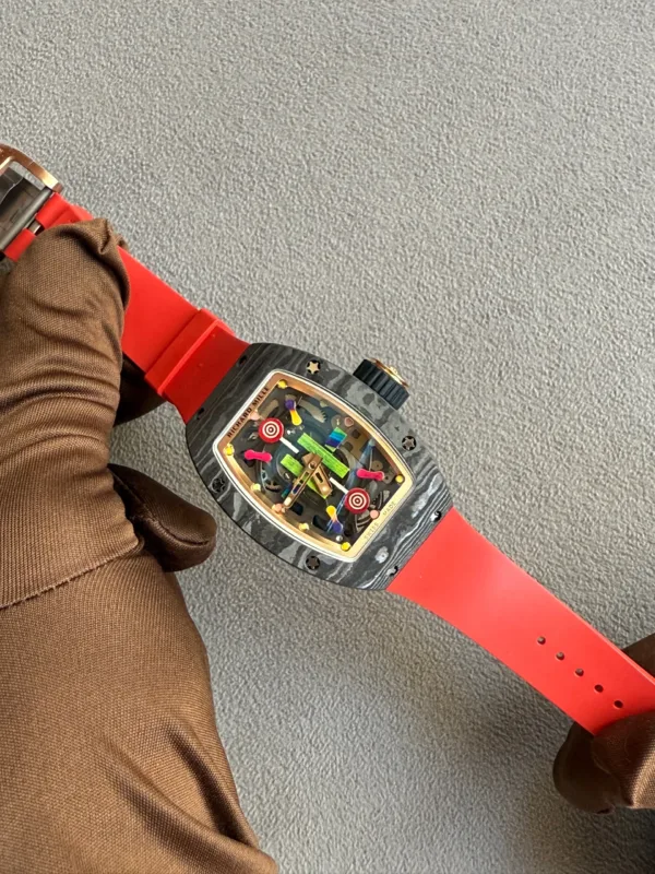 Wristwatch with a bright red strap on a brown-sleeved arm, lying on a gray carpet; see-through colorful dial.