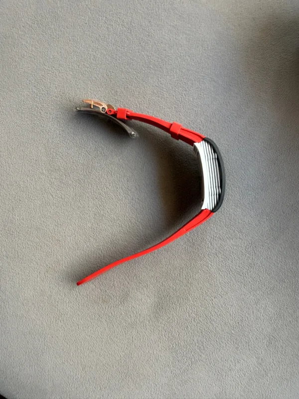 Red wristband with a silver and black watch case on a gray textured surface, showing the band laying flat with a clasp partially visible.
