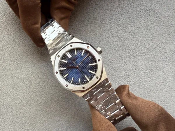 Stainless steel wristwatch with a blue textured dial and an octagonal bezel held by a gloved hand against a gray background.