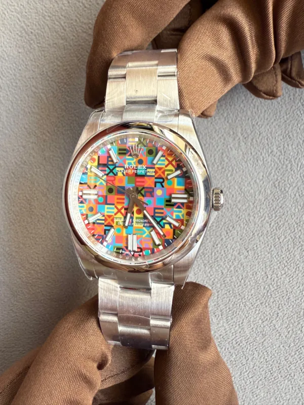 Stainless steel Rolex watch with a multicolored geometric-pattern dial and silver indices, held by a gloved hand against a gray background.
