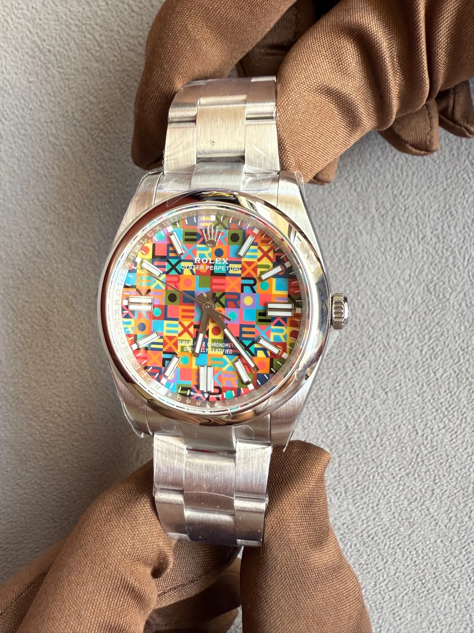 Stainless steel Rolex watch with a multicolored geometric-pattern dial and silver indices, held by a gloved hand against a gray background.