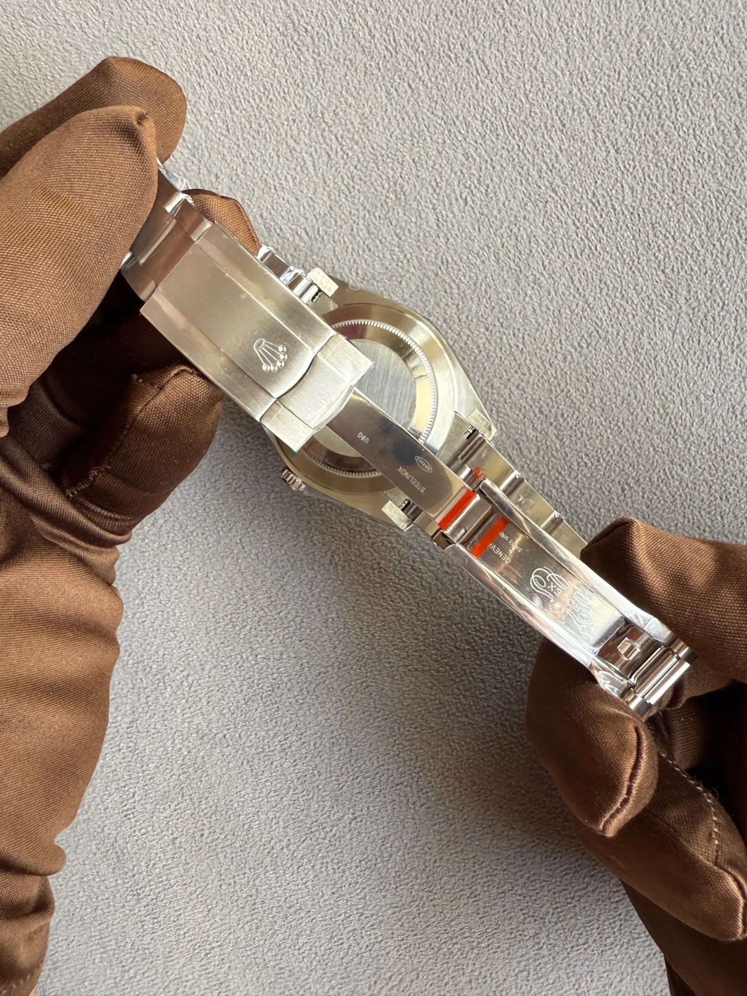 Stainless steel Rolex watch held by brown gloves, showing the clasp and bracelet against a gray textured background