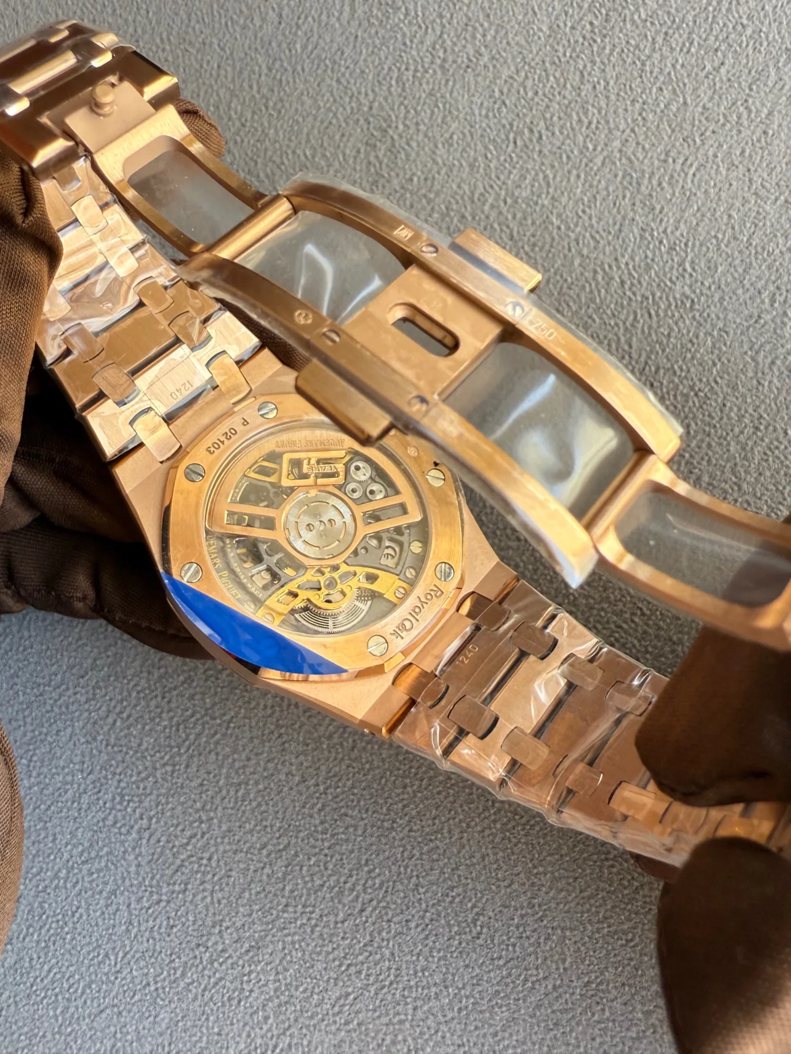 Gold-toned mechanical wristwatch with a transparent case back showing the gears, held by a gloved hand against a blue-gray carpet.
