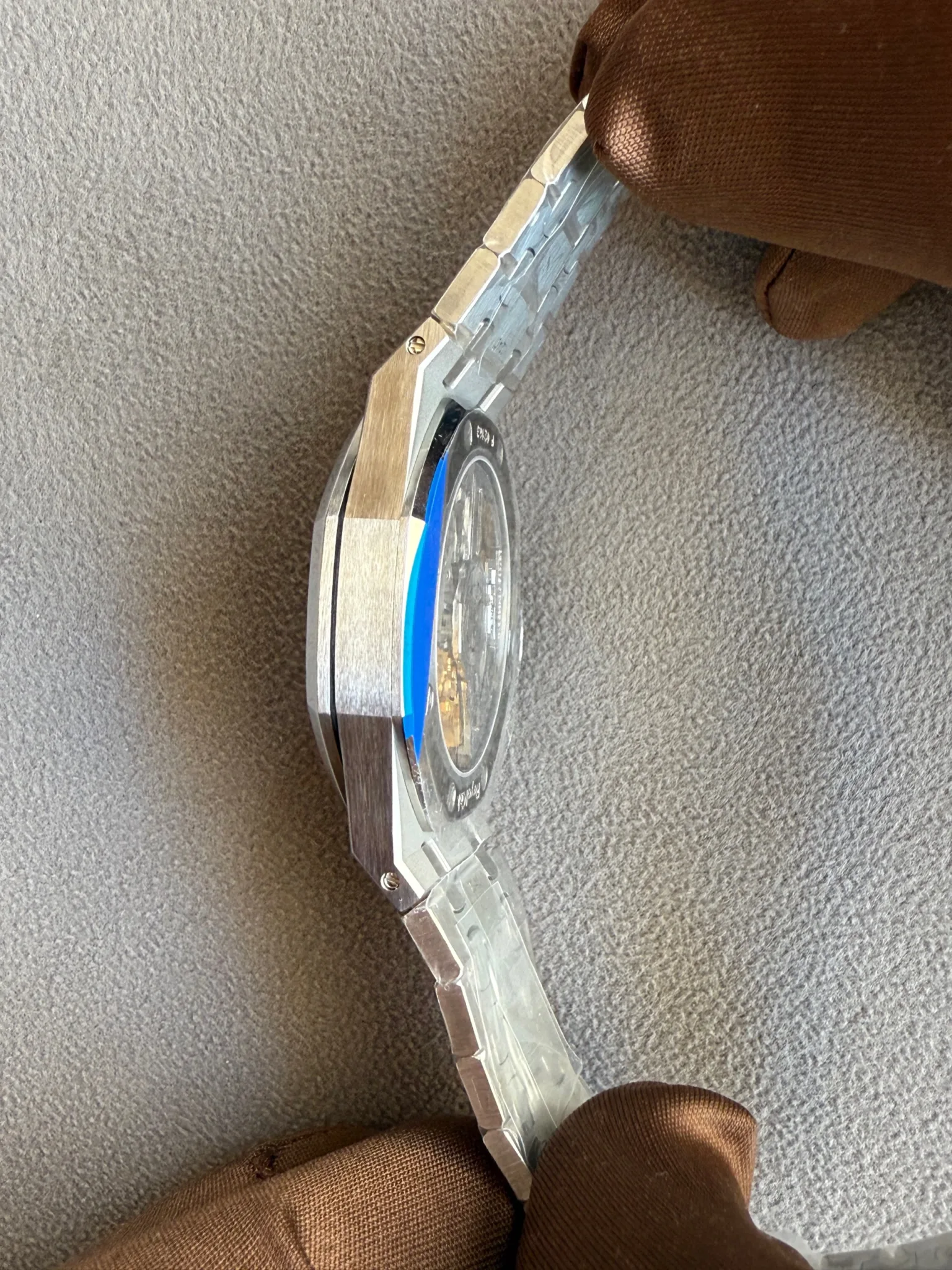 Close-up of a stainless steel watch with a blue protective film on the dial, held by a gloved hand.