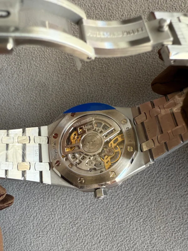 Back view of a stainless steel wristwatch with a transparent case back, revealing gold-toned gears and rotor inside the movement, resting on a textured surface with a metal bracelet partially visible.