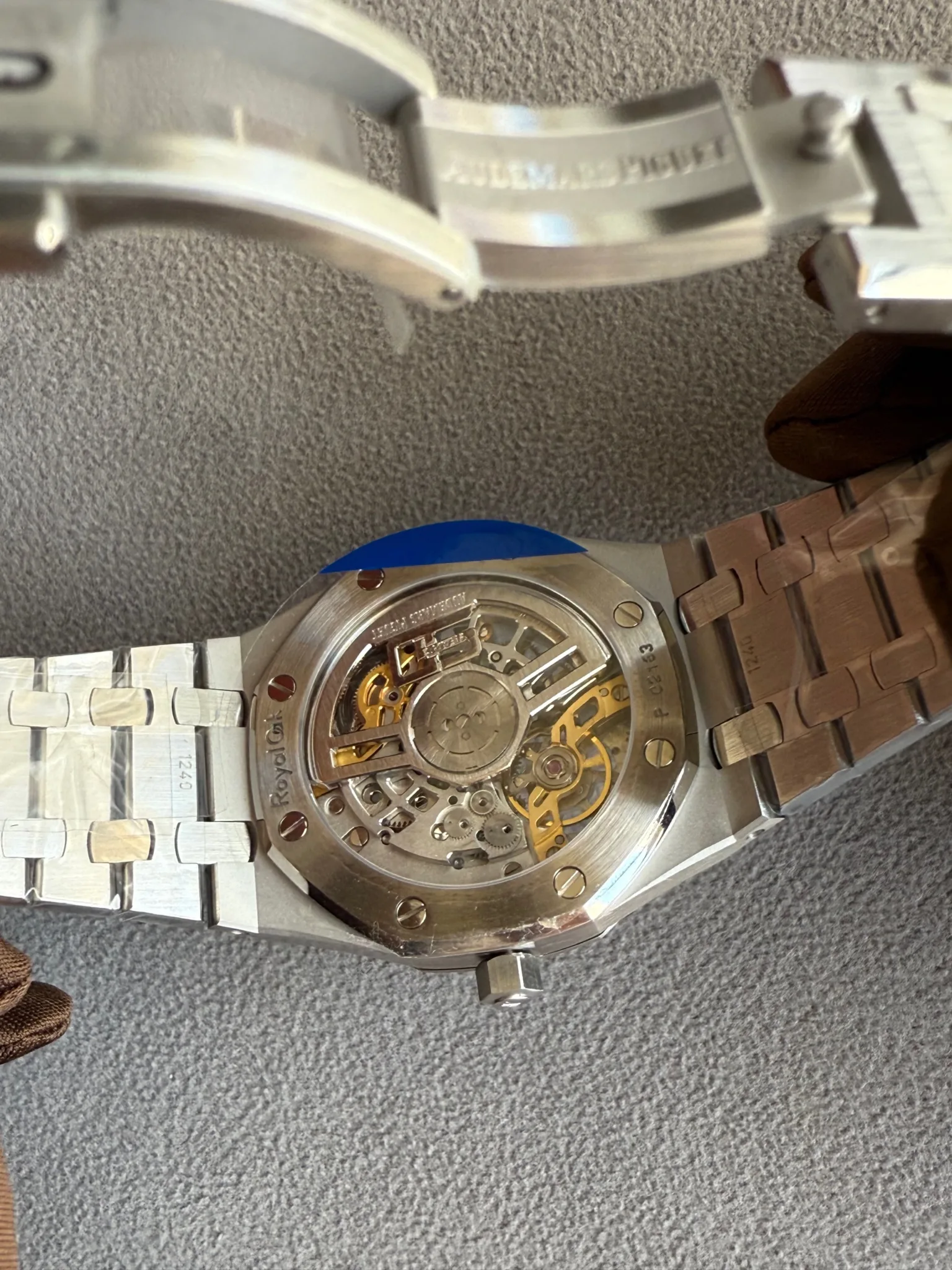Back view of a stainless steel wristwatch with a transparent case back, revealing gold-toned gears and rotor inside the movement, resting on a textured surface with a metal bracelet partially visible.