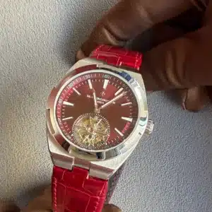 Close-up of a luxury stainless steel wristwatch with a red dial and red crocodile-texture strap, held by a gloved hand against a gray fabric background, showing an open-tourbillon mechanism at the 6 o'clock position.