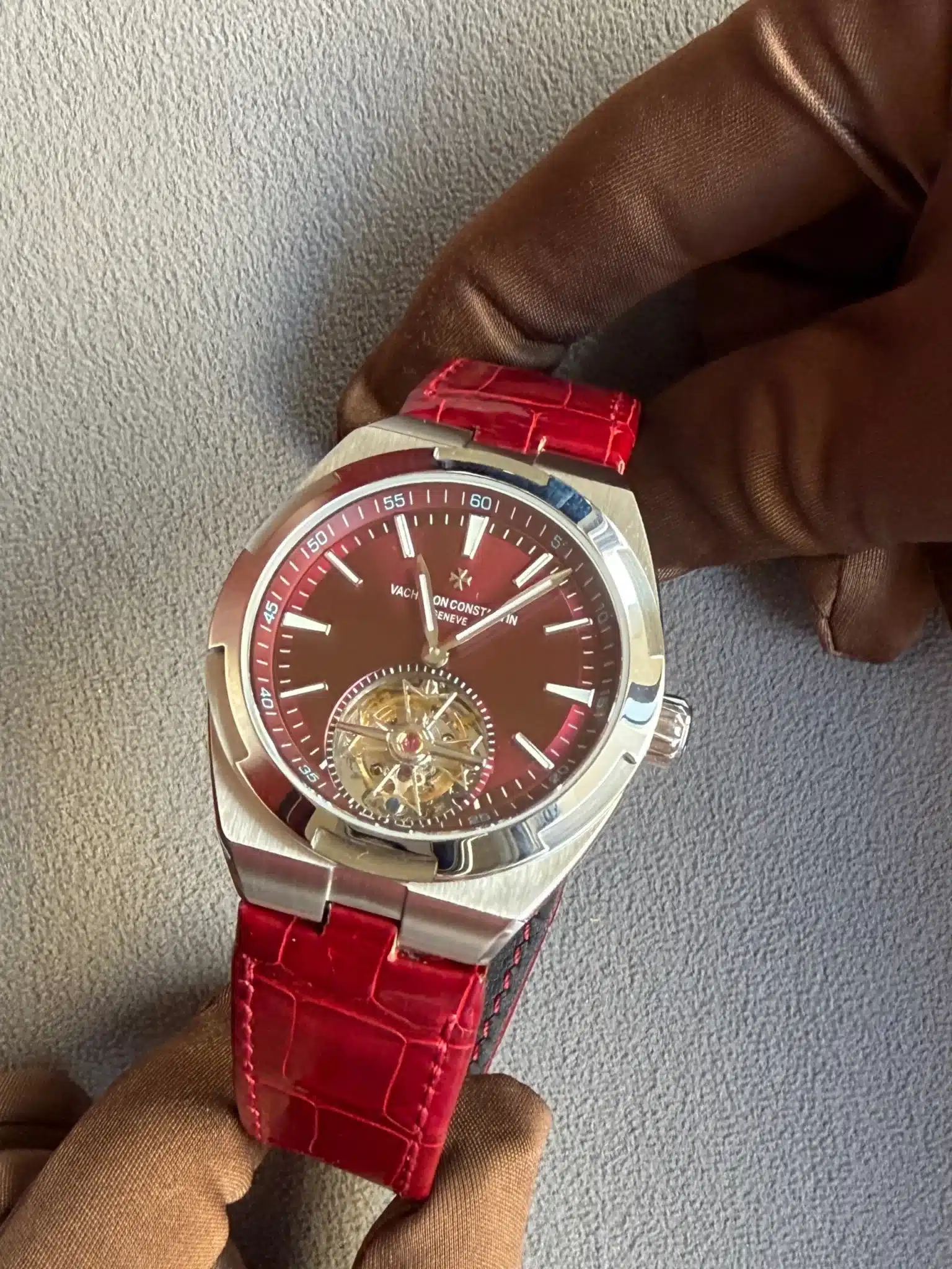 Close-up of a luxury stainless steel wristwatch with a red dial and red crocodile-texture strap, held by a gloved hand against a gray fabric background, showing an open-tourbillon mechanism at the 6 o'clock position.