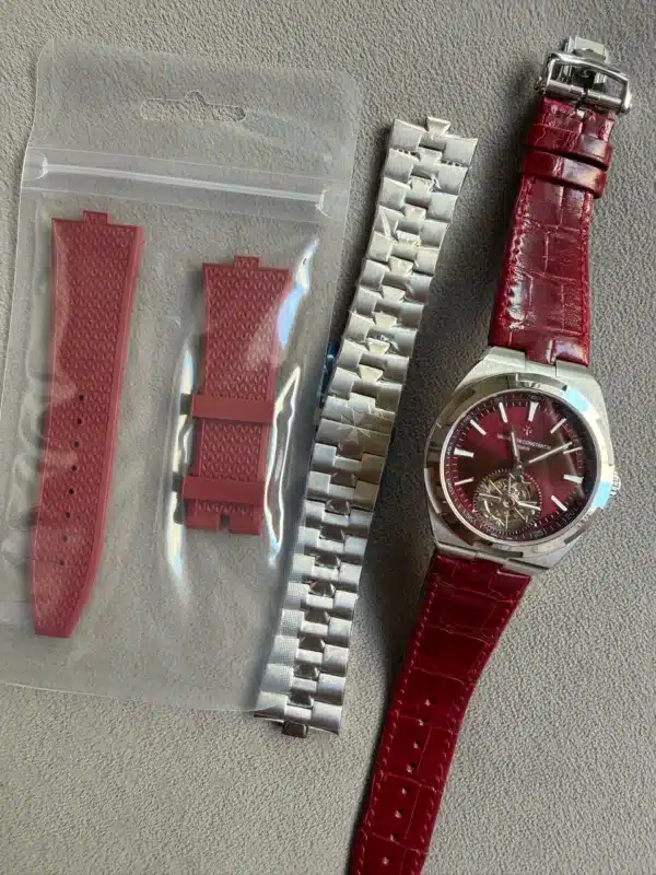Two red rubber watch straps in a clear plastic pouch, alongside a silver metal link bracelet and a red leather strap on a burgundy-dial wristwatch.