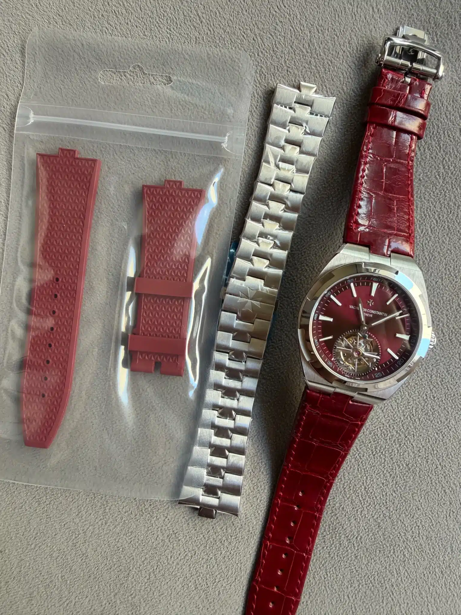 Two red rubber watch straps in a clear plastic pouch, alongside a silver metal link bracelet and a red leather strap on a burgundy-dial wristwatch.